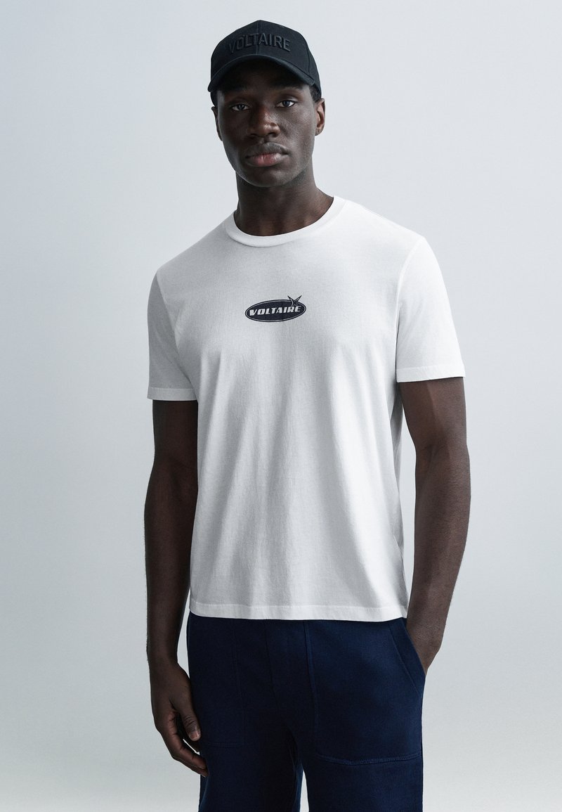 Bílé bavlněné tričko s černým logem "VOLTAIRE" uprostřed, nošené s tmavě modrými kalhotami a sladěnou černou čepicí.