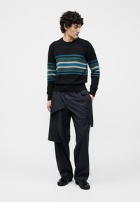 Homme portant un pull noir à rayures horizontales vertes et bleues, un pantalon ample à rayures fines sombres avec un détail en tissu, et des chaussures noires.