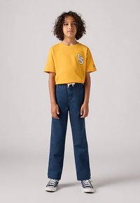 Κίτρινο t-shirt με κοντά μανίκια και λευκό λογότυπο στο στήθος, συνδυασμένο με μπλε τζιν παντελόνι ίσιας γραμμής και ναυτικό μπλε αθλητικά παπούτσια.