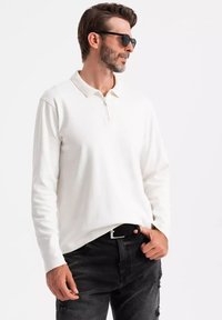 Weißes langärmliges Polohemd mit Reißverschlusskragen, aus weichem Stoff, kombiniert mit schwarzen, ausgewaschenen Jeans und Sonnenbrille. Lässiges, modernes Design.