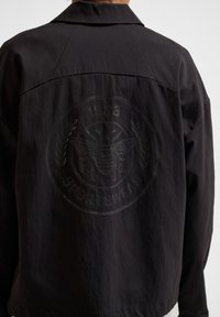 Svart sportjacka i tyg med matt finish, med en texturerad logga med ett bi och lagerkransdesign på baksidan.