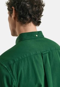 Groene shirt met kraag, voorzien van een drukknoopsluiting en een plooi achterop. De stof lijkt glad te zijn, wat zorgt voor een nette, gestructureerde uitstraling.