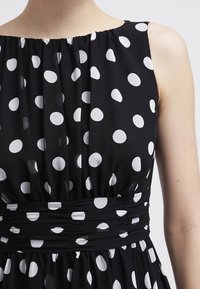 Robe noire à pois blancs, avec un décolleté froncé, un corsage ajusté et une taille élastique pour une jupe fluide.