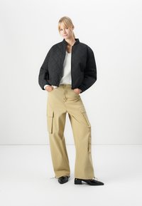 Jeune femme aux cheveux blonds portant une veste matelassée noire, un pull blanc, un pantalon cargo large beige et des chaussures noires à petits talons.