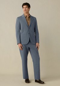 Completo a righe blu chiaro con giacca monopetto, tasche a soffietto e pantaloni abbinati. Indossato sopra una camicia marrone con colletto e scarpe nere.