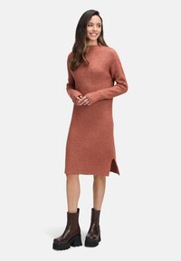 Vrouw in een roestkleurige geribbelde midi-jurk met lange mouwen en bruine chunky haklaarzen, staand en glimlachend.