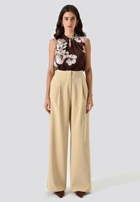Donna che indossa una blusa floreale marrone senza maniche, infilarla in pantaloni a vita alta beige con gamba larga e scarpe a punta marroni.