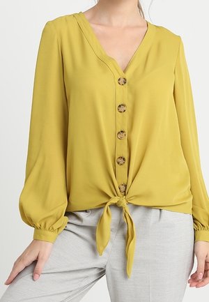 Vrouw draagt een mosterdgele blouse met lange mouwen, houten knopen en een strik aan de voorkant, gecombineerd met een lichtgrijze broek, tegen een effen achtergrond.
