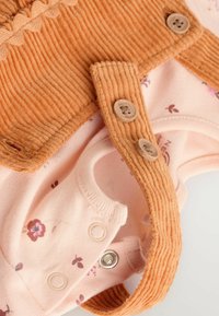 La tenue d'un enfant présente un haut en velours côtelé orange brûlé à côtes avec des bords festonnés et des boutons décoratifs, superposé à un body floral rose clair.