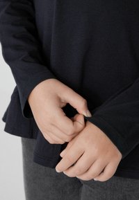 Námornická košile s dlouhým rukávem vyrobená z jemné bavlny, s kulatým výstřihem a nabráním na lemu. Ruce držící košili, zdůrazňující texturu rukávu.