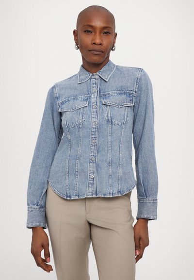 Lyseblå denimskjorte med lange ermer, knappet front, to brystlommer og buet hem. Teksturert med synlige stingdetaljer.
