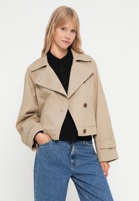 Veste beige courte avec un col cranté, deux boutons marron et des poignets larges, portée sur une chemise noire et associée à un jean bleu.