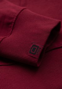 Gros plan sur un tissu côtelé bordeaux plié avec un patch logo rectangulaire noir et rouge cousu près du bord.