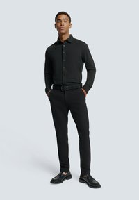 Zwarte blouse met lange mouwen en een getailleerde pasvorm, gecombineerd met zwarte slim-fit broek en zwarte instappers, voor een lichtgrijze achtergrond.