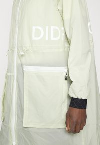 Veste beige clair avec fermeture éclair devant, grande poche rectangulaire zippée, poignet élastique et texte blanc partiel "DIDE" sur la poitrine.