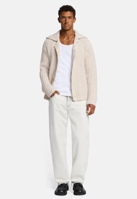 Cardigan beige a maglia grossa con ampio colletto, bottoni frontali e polsini a coste abbinato a pantaloni bianchi e scarpe nere.