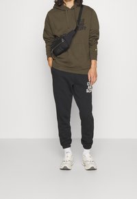 Zielona bluza z kapturem z przednią kieszenią i logo, w zestawie z czarnymi spodniami dresowymi z białymi grafikami; jasnokolorowe sneakersy z teksturowanymi akcentami.