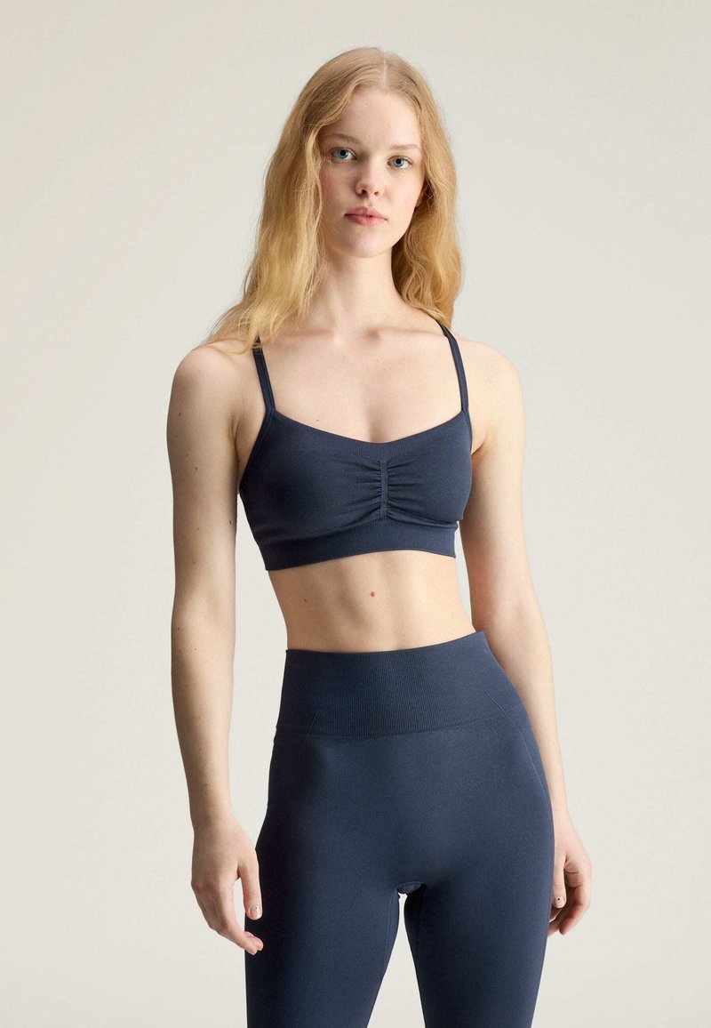Young woman wearing navy blue fitted sports bra and high-waisted leggings, standing with a neutral expression against a plain background.