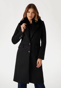 Cappotto lungo in misto lana nero con colletto in pelliccia sintetica, design a bottoni singoli, due tasche frontali e dettagli con bottoni argentati.