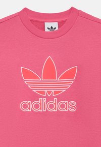 Sweatshirt rosa com gola redonda, textura suave e logo branco da Adidas com emblema de três folhas. Barra reta com mangas curtas.