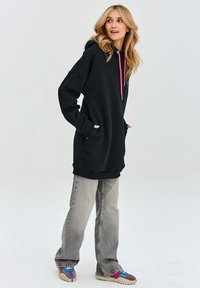 Černá oversized mikina s růžovými tkaničkami, klokankovými kapsami a žebrovanými manžetami, doplněná o světle šedé zvonové džíny a barevné tenisky.