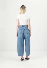 Mujer con cabello rubio largo que lleva una blusa blanca de manga corta, jeans anchos de tiro alto en color azul y zapatos de tacón negros, de pie y mirando hacia atrás.