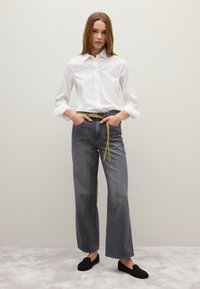 Witte overhemd met lange mouwen, gecombineerd met losse, donkergrijze jeans en een felgele riem. Zwarte instap schoenen maken de outfit compleet.