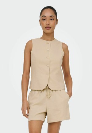 Kvinde iført beige ærmeløs vest med knapper og shorts med snøre, stående foran en ensfarvet lys baggrund.