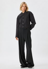 Mustassa lyhyessä takissa on pyöreä pääntie ja viisi nappia, ja se on yhdistetty leveälahkeisiin mustiin housuihin, molemmat sileistä kankaista valmistetut.