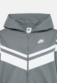 Siva Nike dukserica sa patentnim zatvaračem, s bijelim chevron prugama preko prsnog dijela i Nike logom s lijeve strane.