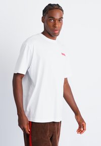 Hvid bomulds T-shirt med korte ærmer, rund halsudskæring og et lille rødt logo på brystet. Kombination med brune bukser og sat mod en ensfarvet baggrund.