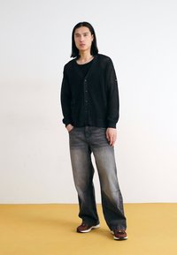 Svart, stickad kofta med V-ringning och knappar, kombinerad med bleknade grå vida jeans och mörka sneakers på ett gult golv.