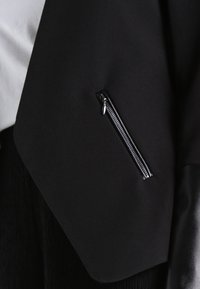 Černá bunda s hladkou látkou, která má na přední straně kapsu na zip a kožené doplňky na rukávech.