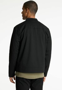 Zwarte jas met een eenvoudig ontwerp, met een hoge kraag en een textuurstof. Zichtbare stiksels en gelaagd met een groene shirt eronder.