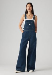 Mørkeblå denim overalls med vide ben, frontlomme, justerbare stropper og sølvhardware. Brukt over en hvit tanktop.