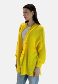 Gul stickad cardigan med en lös passform, med rund hals, stora gyllene knappar och ribbade muddar. Bärs över en grå topp.
