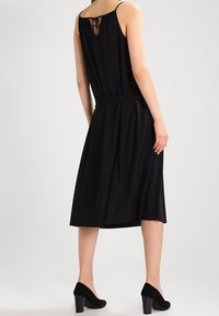 Schwarzes ärmelloses Kleid mit tailliertem Bund, das einen mittellangen Rock und ein offenes Rückendesign mit einer Schleife aufweist. Kombiniert mit schwarzen High Heels.