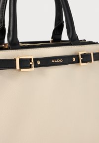 Beige Lederhandtasche mit schwarzen Akzenten und Griffen. Hardware in Goldfarbe und ein markierter schwarzer Riemen. Strukturierte Oberfläche. Reißverschlussverschluss.
