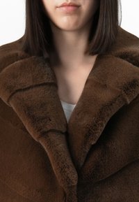 Donna che indossa un cappotto di pelliccia marrone scuro, sopra una maglietta bianca, con i capelli scuri e lisci visibili sulle spalle.