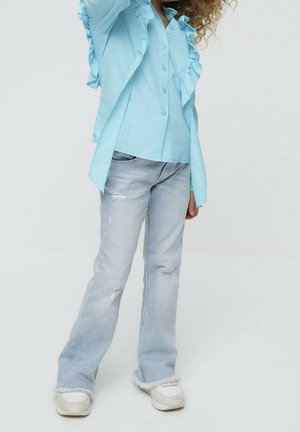 Niño con camisa azul claro de mangas con volantes, jeans desgastados de lavado claro y zapatillas blancas, de pie contra un fondo blanco liso.