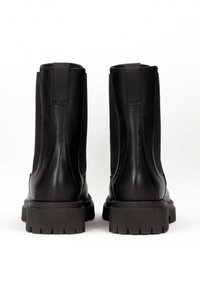 Para czarnych skórzanych botków za kostkę z grubymi, teksturowanymi gumowymi podeszwami, widziana od tyłu na białym tle.