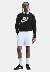Sudadera negra de Nike con logo blanco, pantalones cortos blancos, zapatillas negras y calcetines blancos. El modelo está de pie con las manos en los bolsillos frente a un fondo neutro.