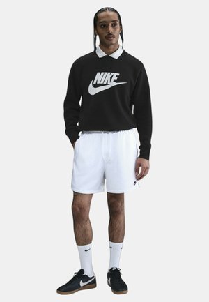 Felpa nera Nike con logo bianco, pantaloni corti bianchi, sneakers nere e calze bianche. Il modello sta in piedi con le mani in tasca contro uno sfondo neutro.