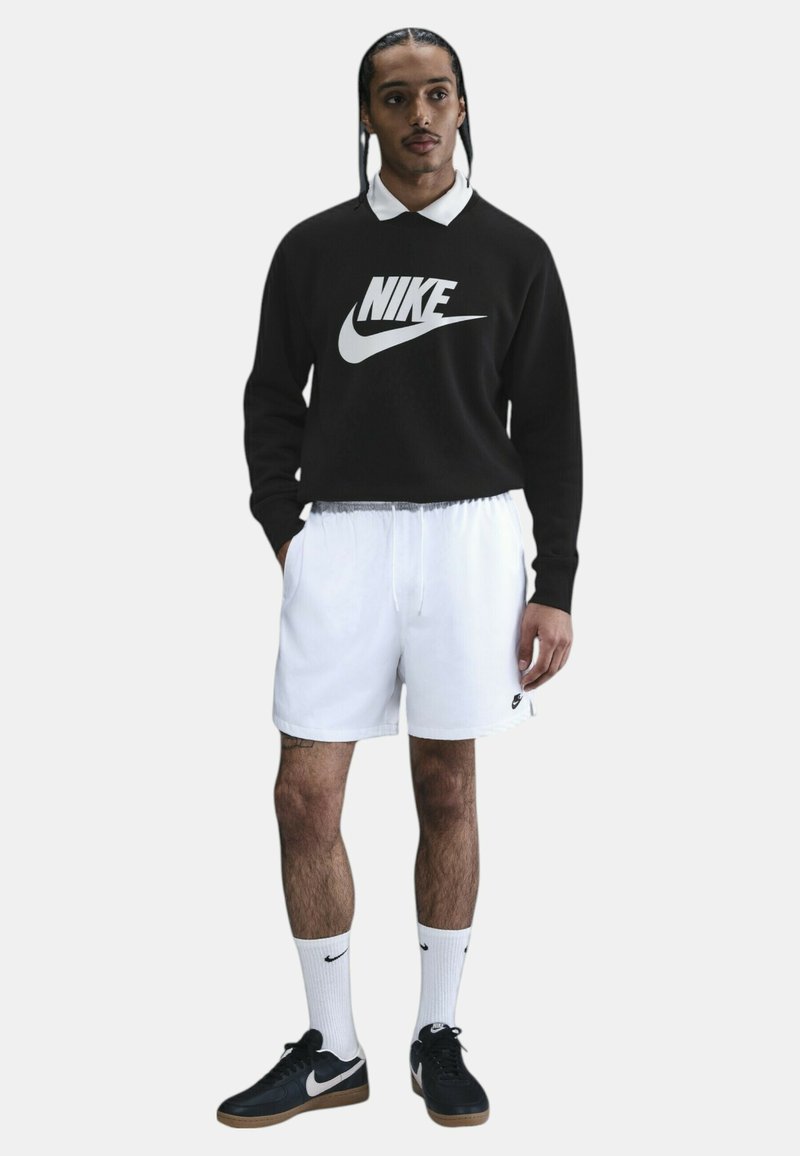 Sudadera negra de Nike con logo blanco, pantalones cortos blancos, zapatillas negras y calcetines blancos. El modelo está de pie con las manos en los bolsillos frente a un fondo neutro.