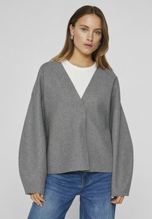Grå oversized tröja med V-ringning ochvida ärmar, tillverkad av ett mjukt, texturerat tyg, lager på lager över en vit skjorta. Blå jeans fulländar outfiten.