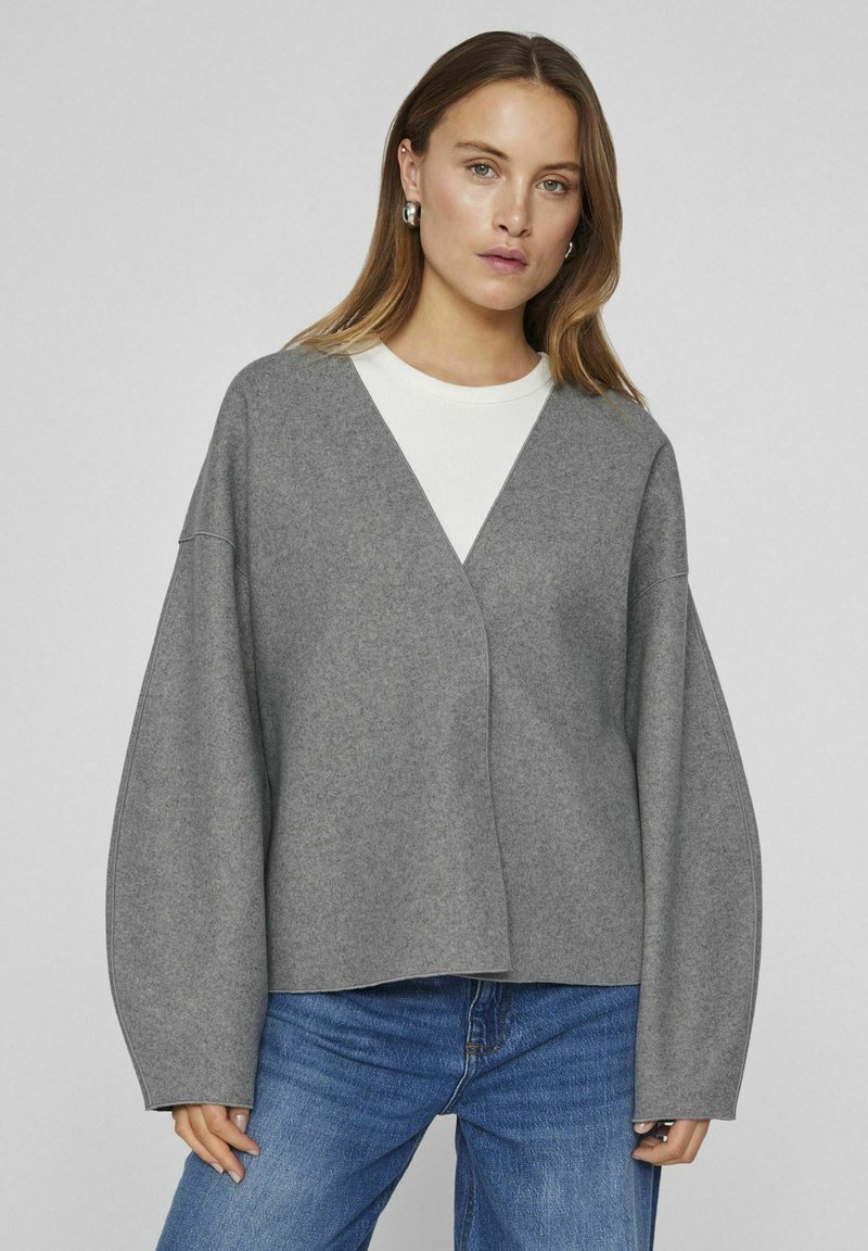 Grå oversized tröja med V-ringning ochvida ärmar, tillverkad av ett mjukt, texturerat tyg, lager på lager över en vit skjorta. Blå jeans fulländar outfiten.
