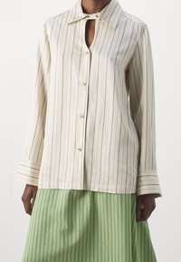 Camicia con bottoni, colletto, apertura a occhio di chiave, tessuto crema con sottili righe marroni e maniche a campana. Indossata con una gonna plissettata verde.