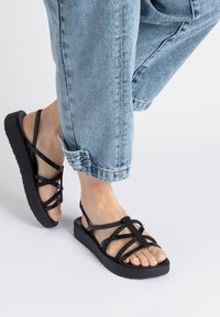 Schwarze Plattform-Sandalen mit überkreuzten Riemen und strukturiertem Sohle. Getragen mit hellblauen, hochgekrempelten Jeans, die die Zehen zeigen.