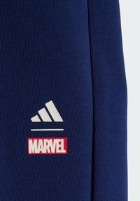 Detailný záber námornícky modrej látky s bielym logom Adidas nad bielym logom Marvel s červeným okrajom.