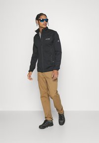 Giacca nera con zip e colletto alto, con logo sul petto e tasca laterale; abbinata a pantaloni cargo beige e scarpe nere.